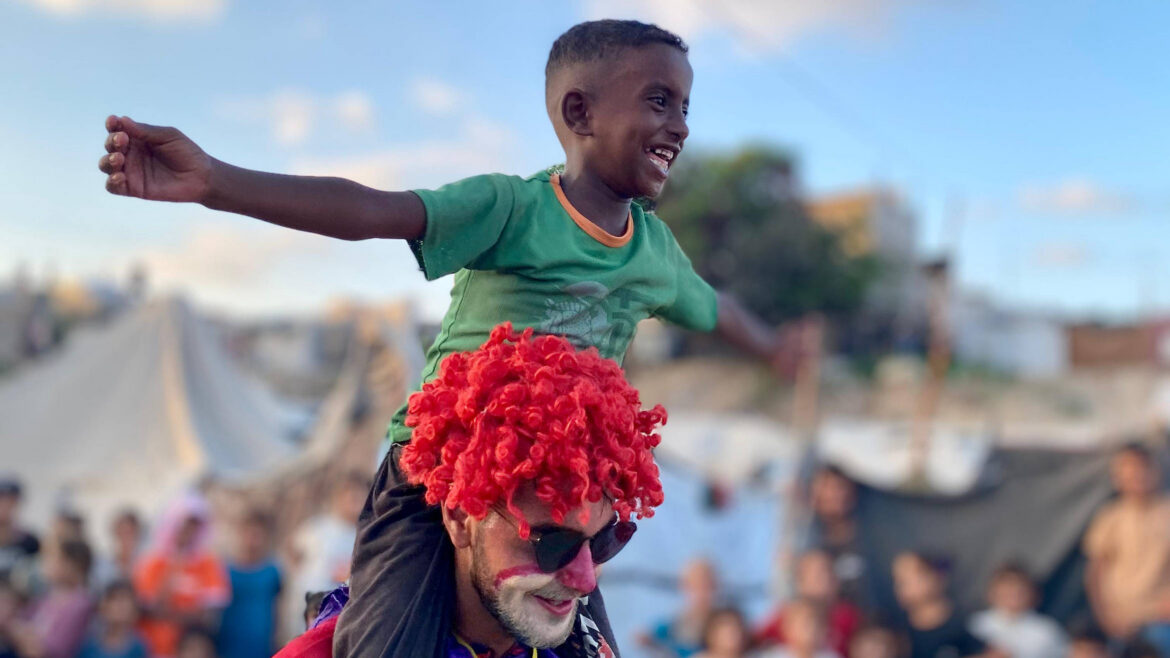 A boy laughs with his arms spread wide while riding on the shoulders of a volunteer dressed as a clown, as a crowd of children watches.