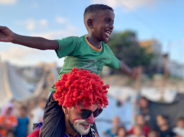 A boy laughs with his arms spread wide while riding on the shoulders of a volunteer dressed as a clown, as a crowd of children watches.